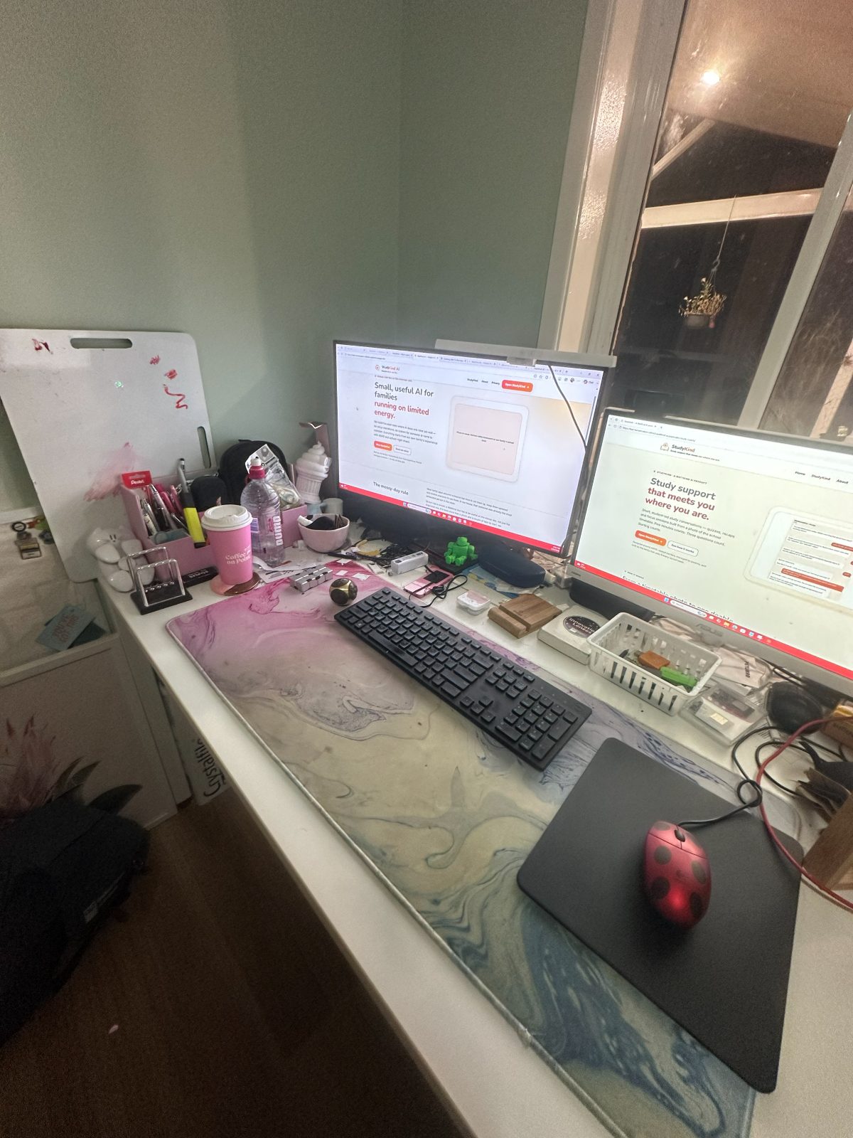 The desk WattKind is built at, on an ordinary evening: two monitors by a dark window, a keyboard on a marbled desk mat, and the everyday clutter around them — pens, a water bottle, a coffee cup, a whiteboard, tangled cables.